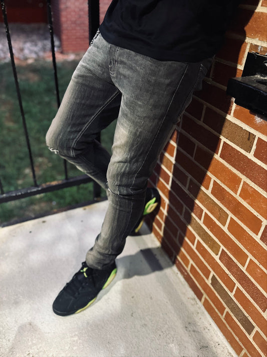 Person wearing gray jeans and black shoes leaning against a brick wall.